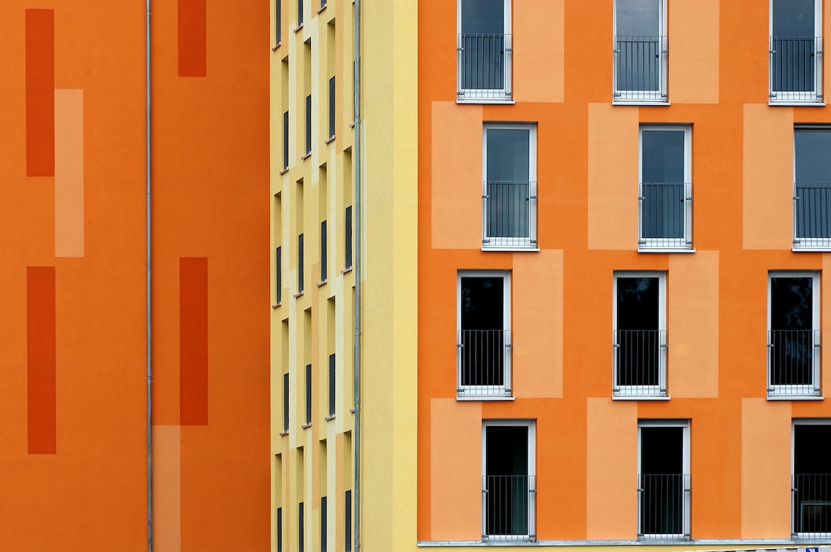 Modern, colorful façade of an apartment building in shades of orange and yellow with regular window fronts — a symbol of the Swiss rental housing market and the claim to a rent reduction as the reference interest rate falls. “>
