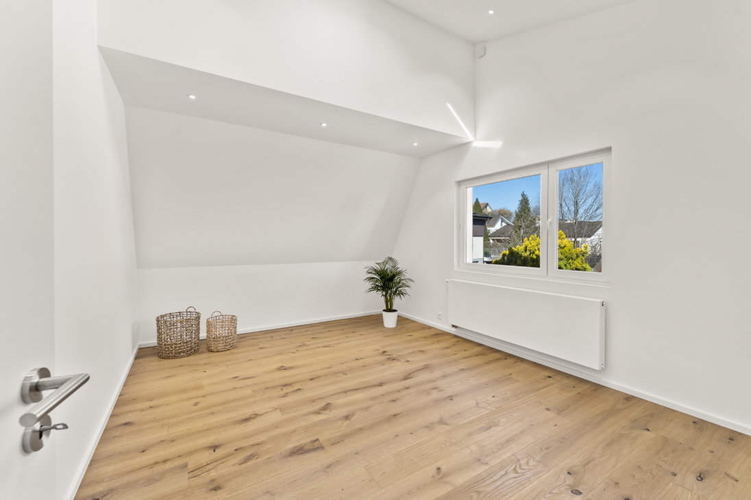 Bedroom with sloped ceiling in a renovated detached house in Zürich featuring oak hardwood flooring, large window with natural light, radiator, and potted plants.