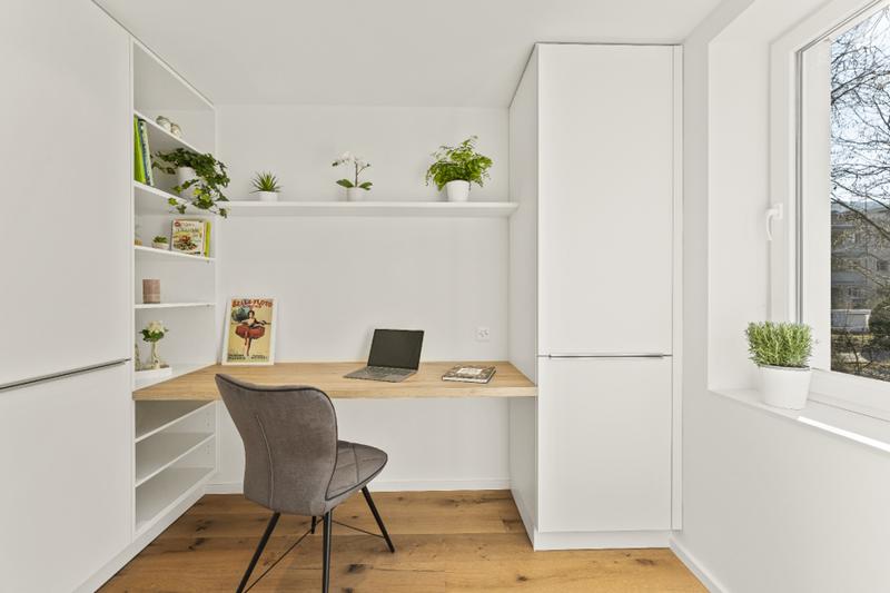 Bright home office in a renovated detached house in Zürich with built-in white shelving, oak desk, grey upholstered chair, laptop, plants, and hardwood flooring.