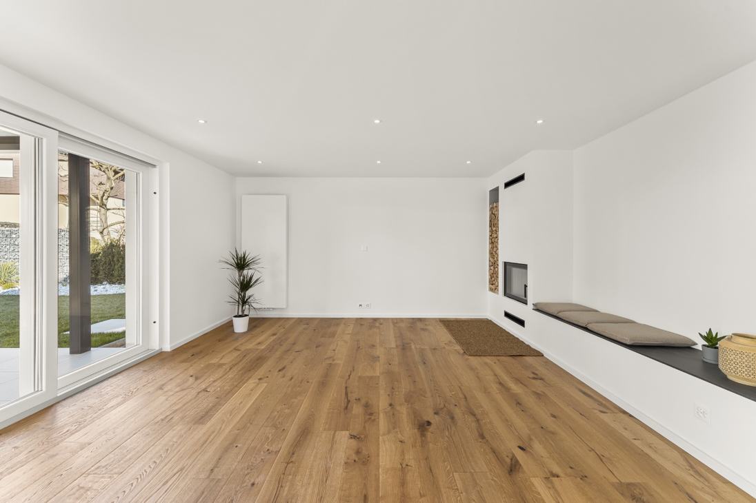 Spacious living room in a renovated detached house in Zürich with built-in fireplace, integrated wood storage, cushioned bench seat, large sliding glass doors to garden, and oak flooring.