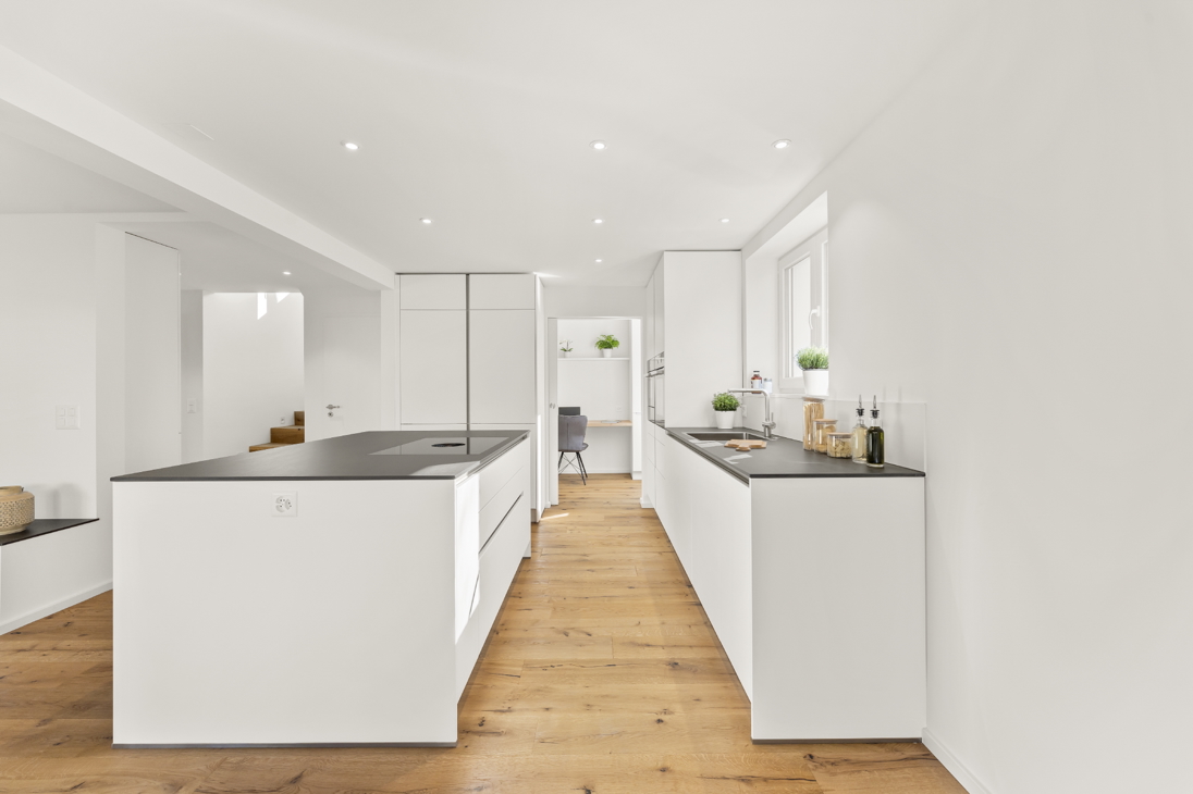 Modern white handleless kitchen in a renovated detached house in Zürich with dark stone countertops, large island with induction hob, oak flooring, and window with greenery.